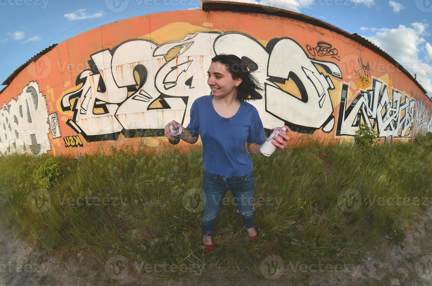 portrait d'une jeune fille émotionnelle aux cheveux noirs et aux piercings. une photo grand angle d'une fille avec des pots de peinture aérosol dans les mains sur un fond de mur de graffitis. un portrait moderne d'un objectif fisheye