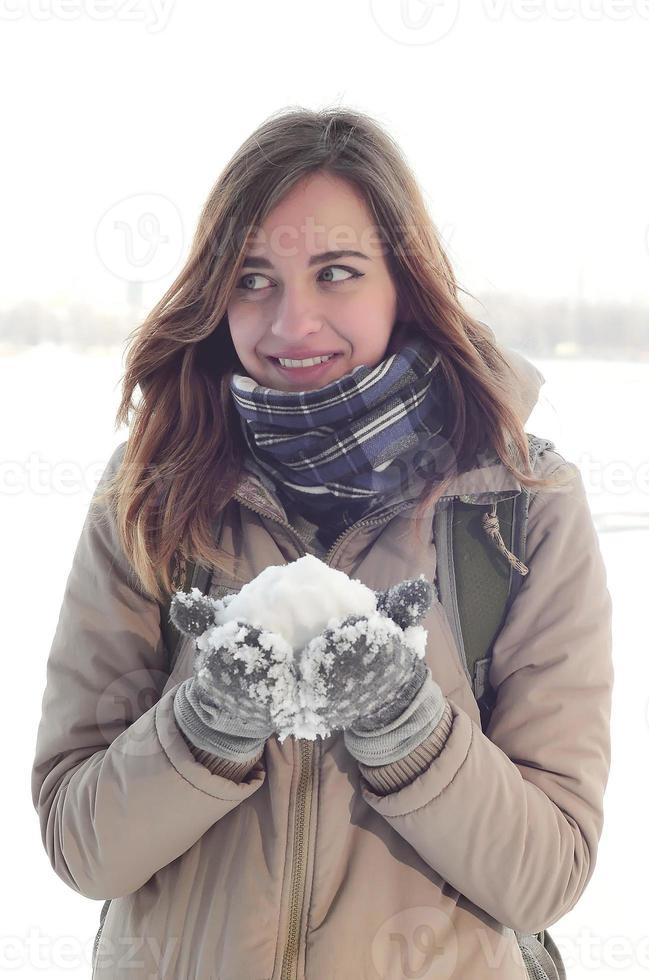 une jeune et joyeuse fille caucasienne dans un manteau marron tient une boule de neige à l'arrière-plan d'une ligne d'horizon entre le ciel et un lac gelé en hiver photo