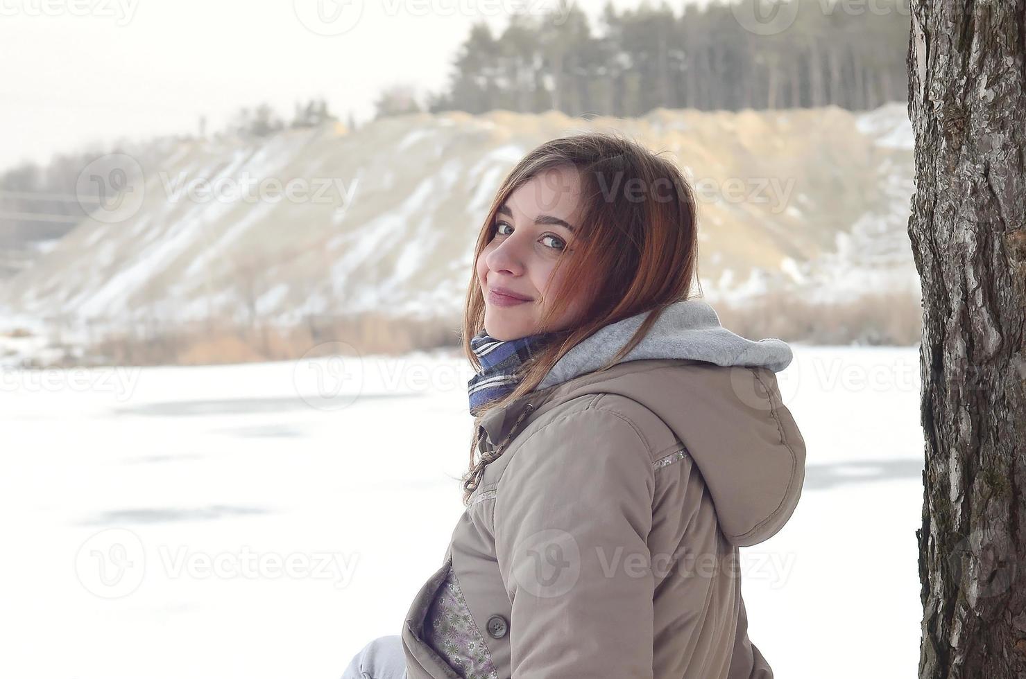 une jeune fille de race blanche dans un manteau marron est assise près d'une falaise à l'arrière-plan d'une ligne d'horizon entre le ciel et un lac gelé en hiver photo