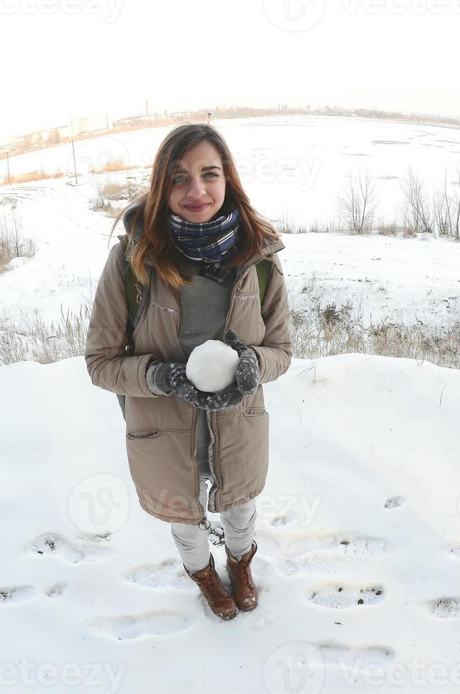 une jeune et joyeuse fille caucasienne en manteau marron tient une boule de neige devant une ligne d'horizon entre le ciel et un lac gelé en hiver. photo fisheye