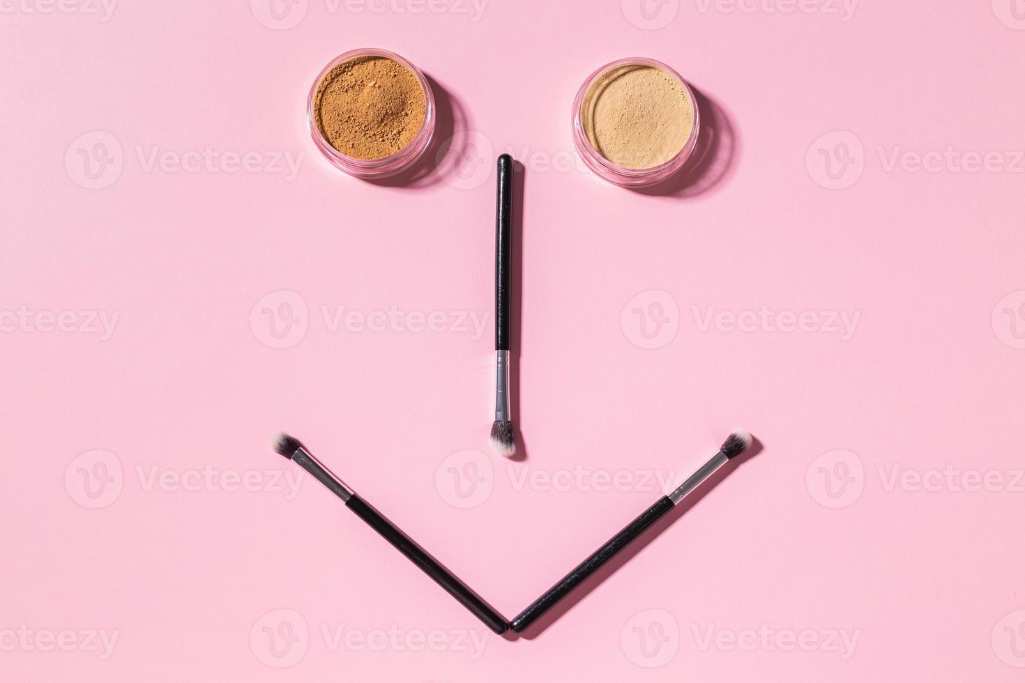 visage amusant avec pinceaux de maquillage et poudre minérale sur fond rose avec espace de copie, vue de dessus. concept d'humour et de beauté cosmétique. photo