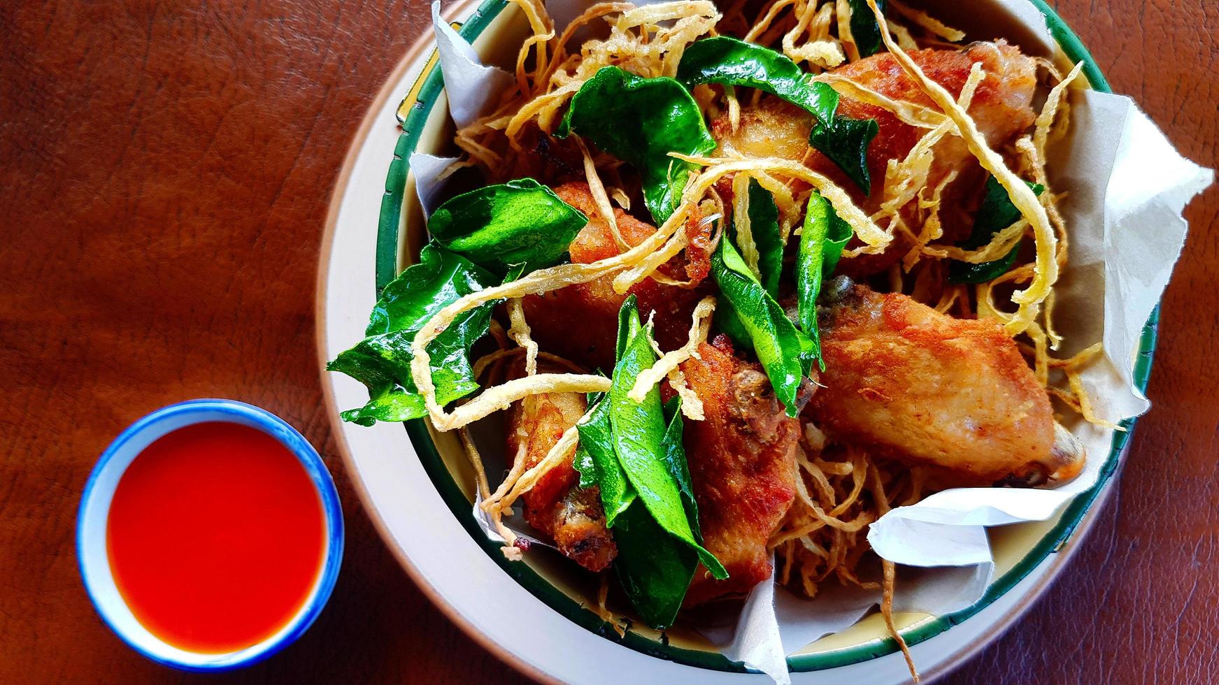 mise à plat d'ailes de poulet frites avec des feuilles de lime kaffir croustillantes et de la papaye râpée sur un bol et de la sauce chili sur une table brune. vue de dessus de la cuisine thaïlandaise avec des herbes et des légumes frits. photo