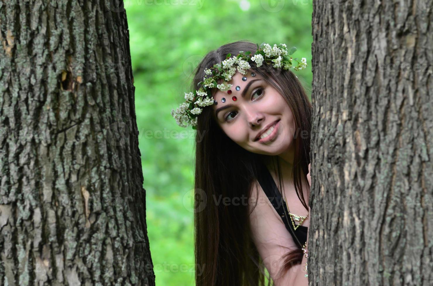 une photo de forêt d'une belle jeune brune d'apparence européenne aux yeux marron foncé et aux grandes lèvres. sur la tête de la jeune fille porte une couronne de fleurs, sur son front des décorations brillantes