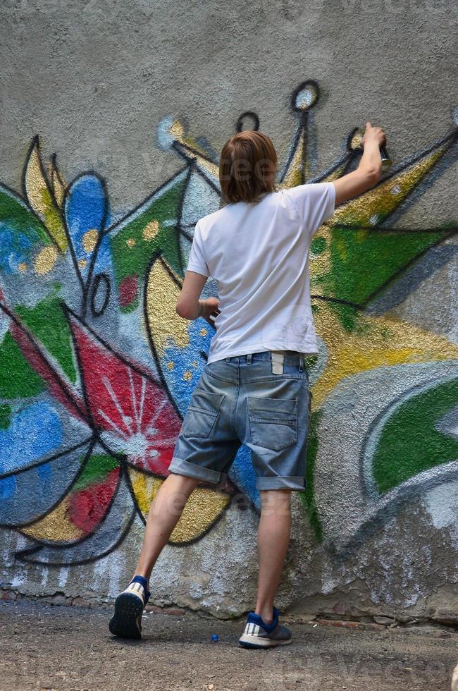 photo d'un jeune homme en short en jean et chemise blanche. le gars dessine sur le mur de graffitis un dessin avec des peintures aérosols de différentes couleurs. le concept de hooliganisme et de dommages aux biens