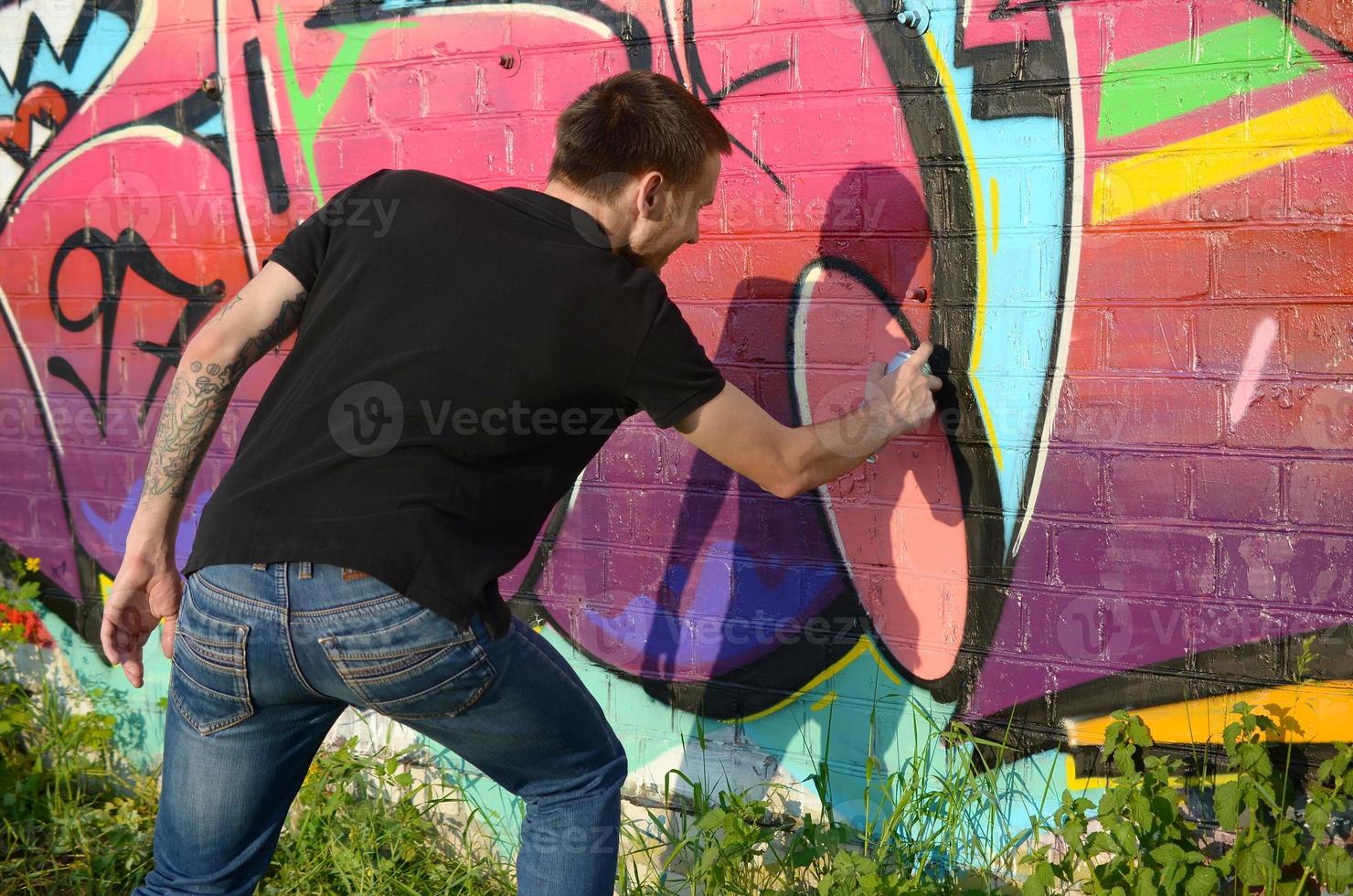 jeune graffeur avec sac à dos et masque à gaz sur son cou peint des graffitis colorés dans des tons roses sur un mur de briques. street art et processus de peinture contemporaine photo