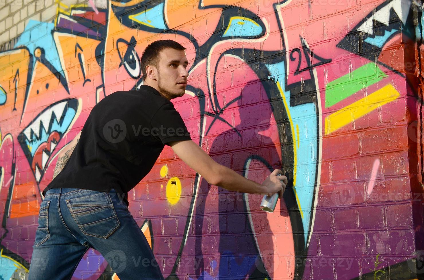 jeune graffeur avec sac à dos et masque à gaz sur son cou peint des graffitis colorés dans des tons roses sur un mur de briques. street art et processus de peinture contemporaine photo