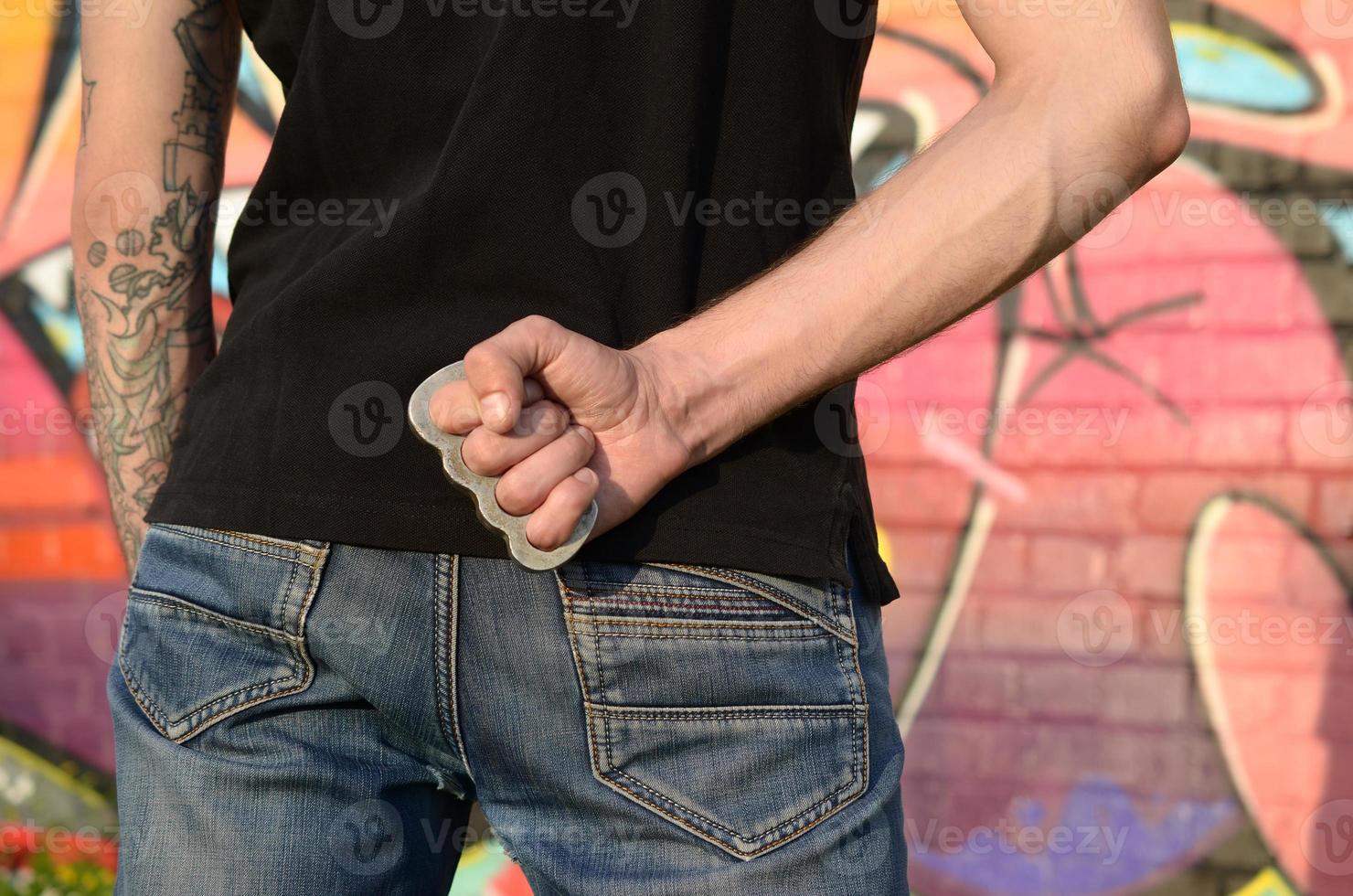 vue arrière du jeune homme de race blanche avec un cnuckle en laiton sur sa main contre le mur de briques du ghetto avec des peintures de graffitis. concept de forces criminelles et accusation d'agression photo