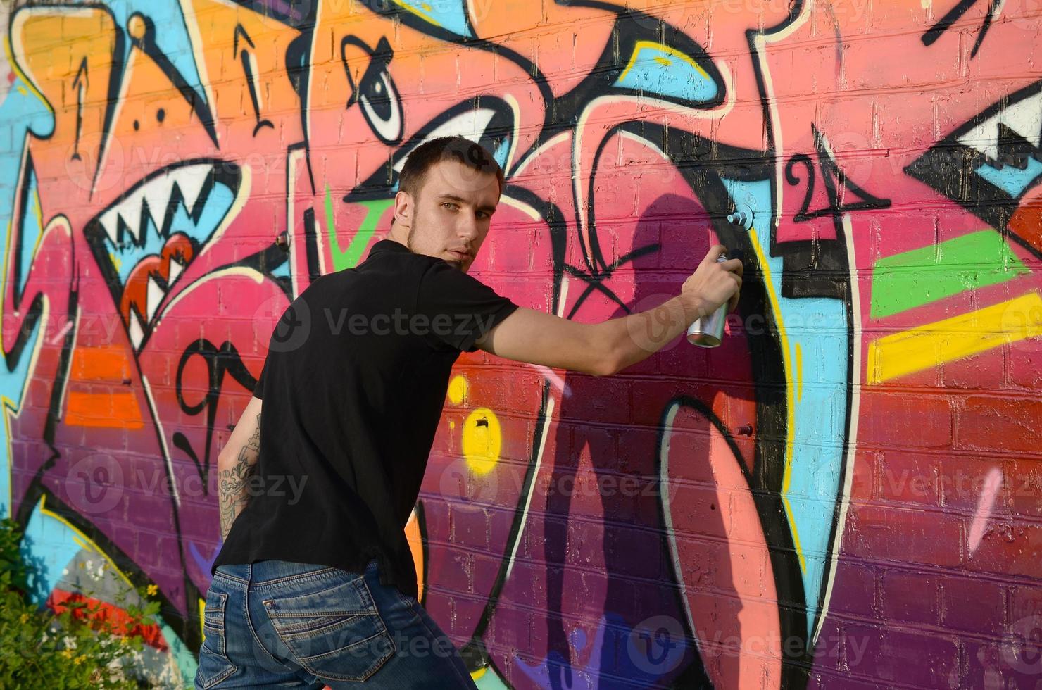 jeune graffeur avec sac à dos et masque à gaz sur son cou peint des graffitis colorés dans des tons roses sur un mur de briques. street art et processus de peinture contemporaine photo