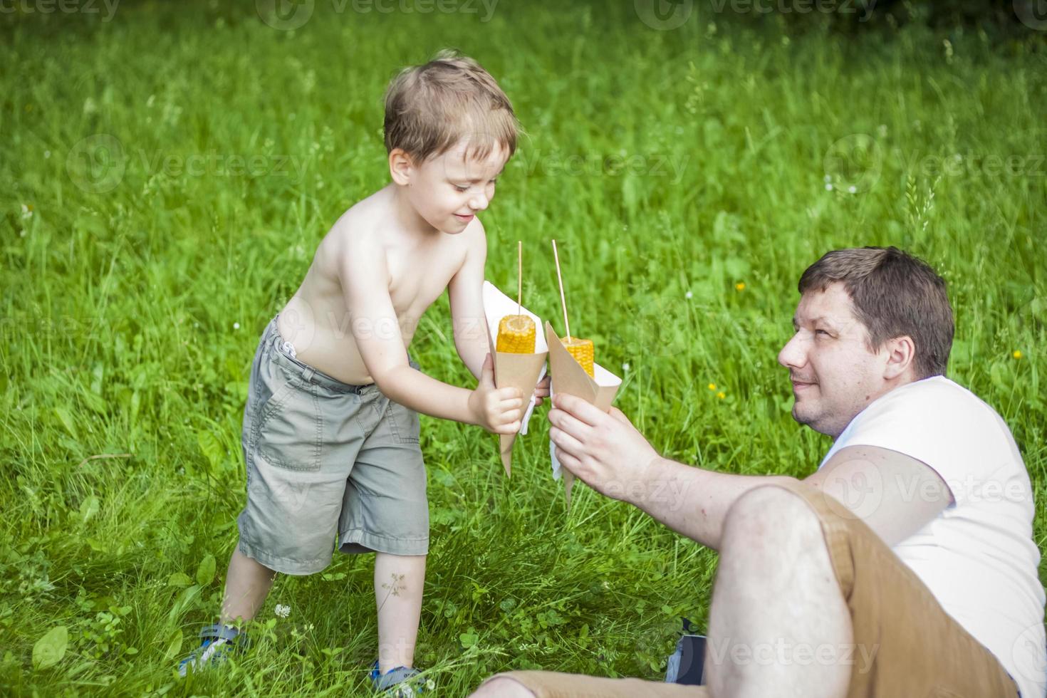 un mignon garçon blond et son père mangent du maïs en été, assis au bord de la rivière sur l'herbe juteuse. drôle d'expression faciale. photo