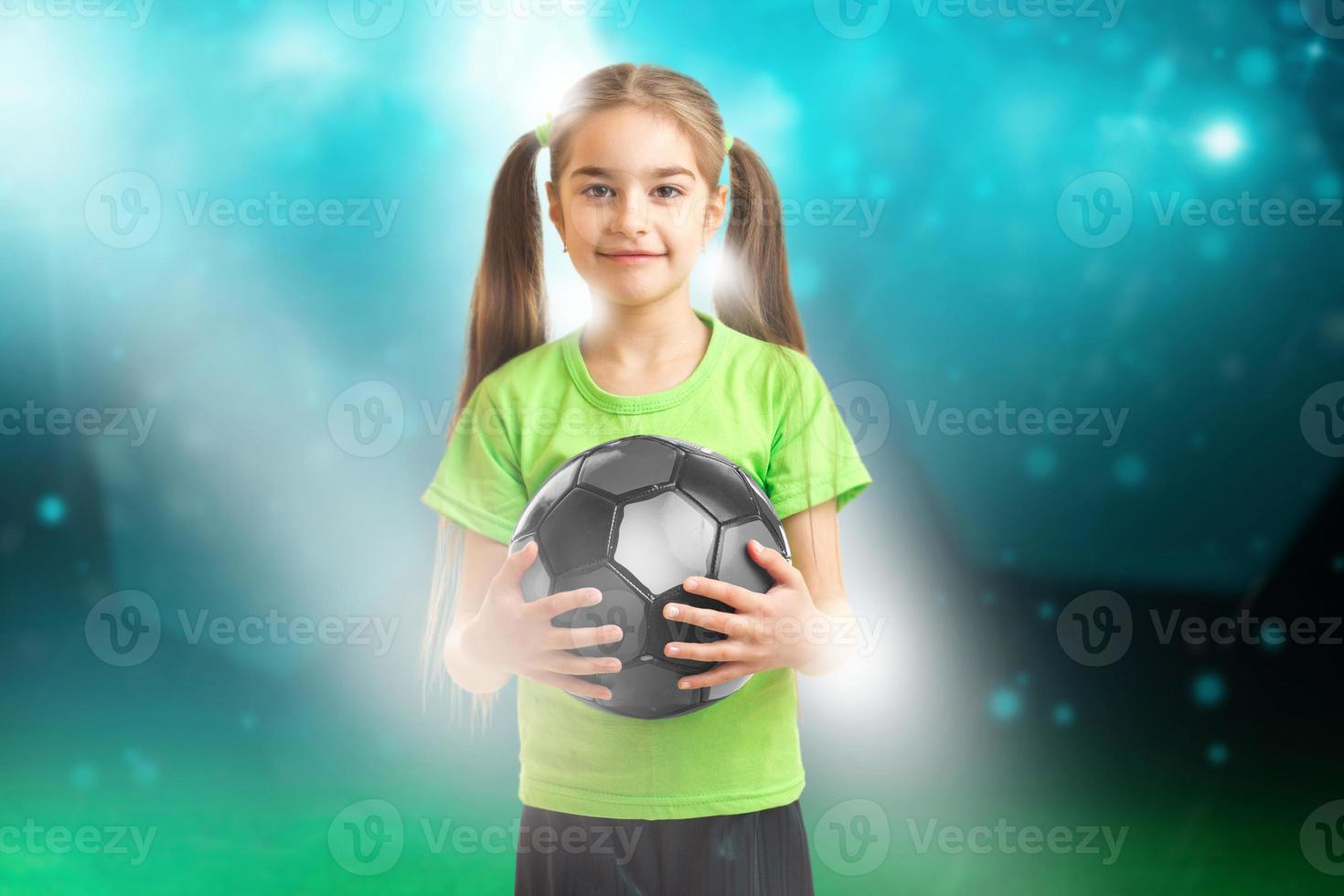 et sourit à la caméra petite fille en chemise verte tenant un ballon de football photo