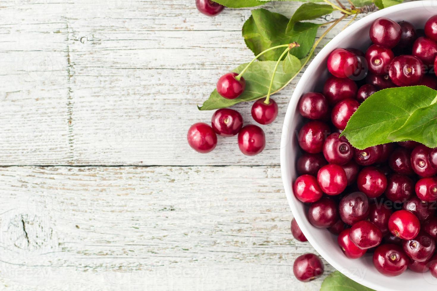 bol blanc de cerises douces fraîches avec des feuilles dans des gouttes d'eau sur fond de bois photo