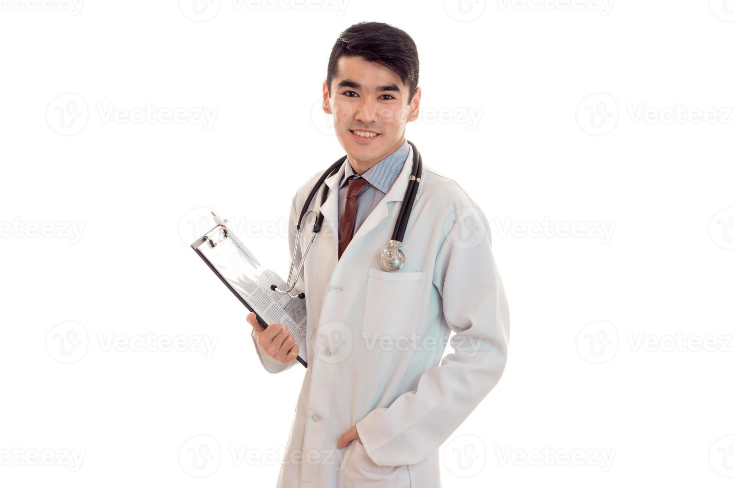 jeune médecin brune élégante en uniforme avec stéthoscope souriant à la caméra et prendre des notes isolées sur fond blanc photo