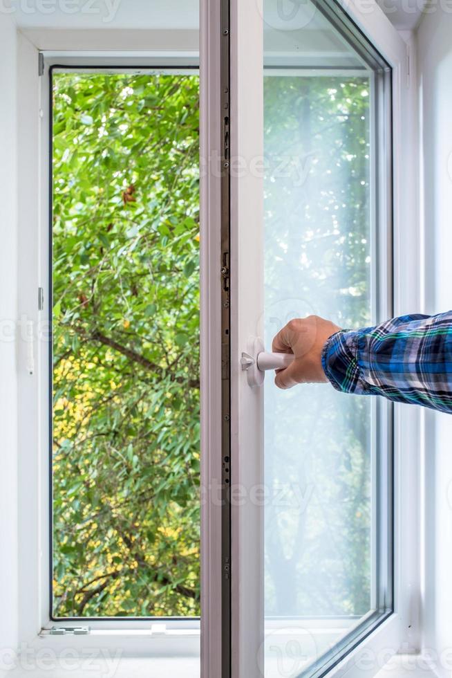 la main de l'homme ouvre une fenêtre en pvc à partir de laquelle vous pouvez voir un arbre dans une zone écologiquement propre de la ville, avec de l'air frais. photo