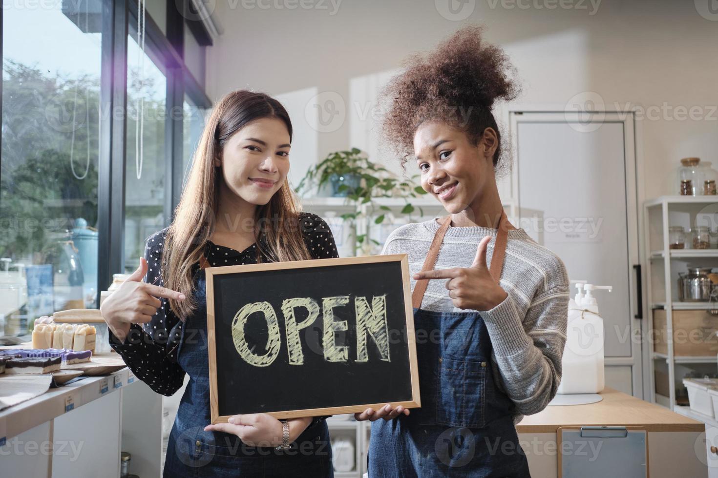 deux jeunes commerçantes montrent un panneau ouvert avec des sourires joyeux dans un magasin de recharge, un travail heureux avec des produits biologiques, des produits d'épicerie zéro déchet, des marchandises respectueuses de l'environnement et des entreprises durables. photo