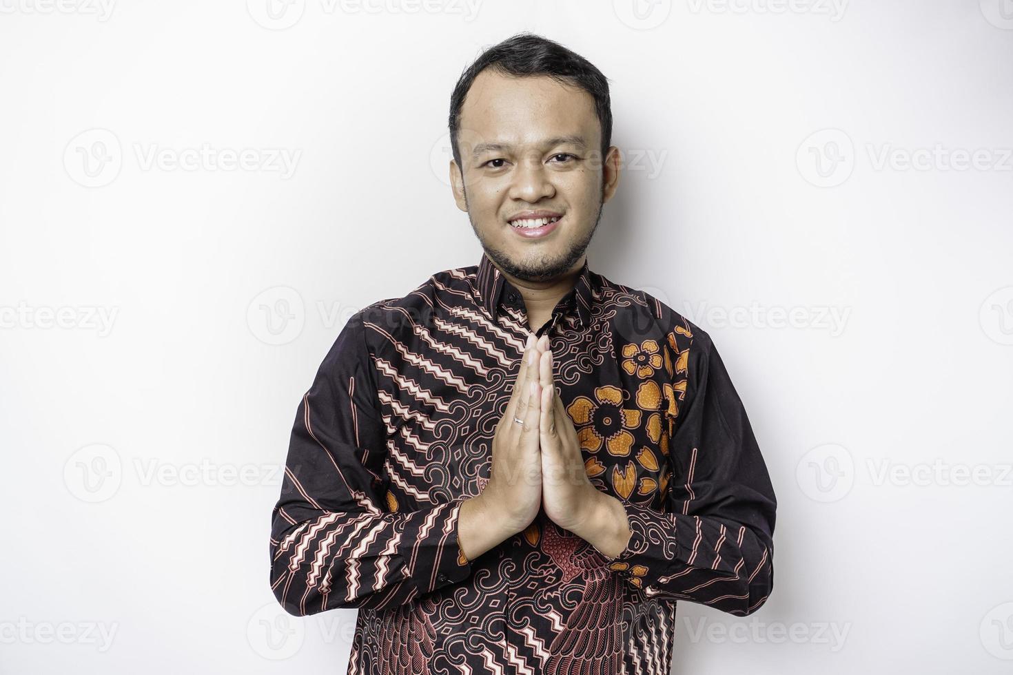 jeune homme asiatique souriant portant une chemise batik, faisant des gestes de salutation traditionnelle isolés sur fond blanc photo