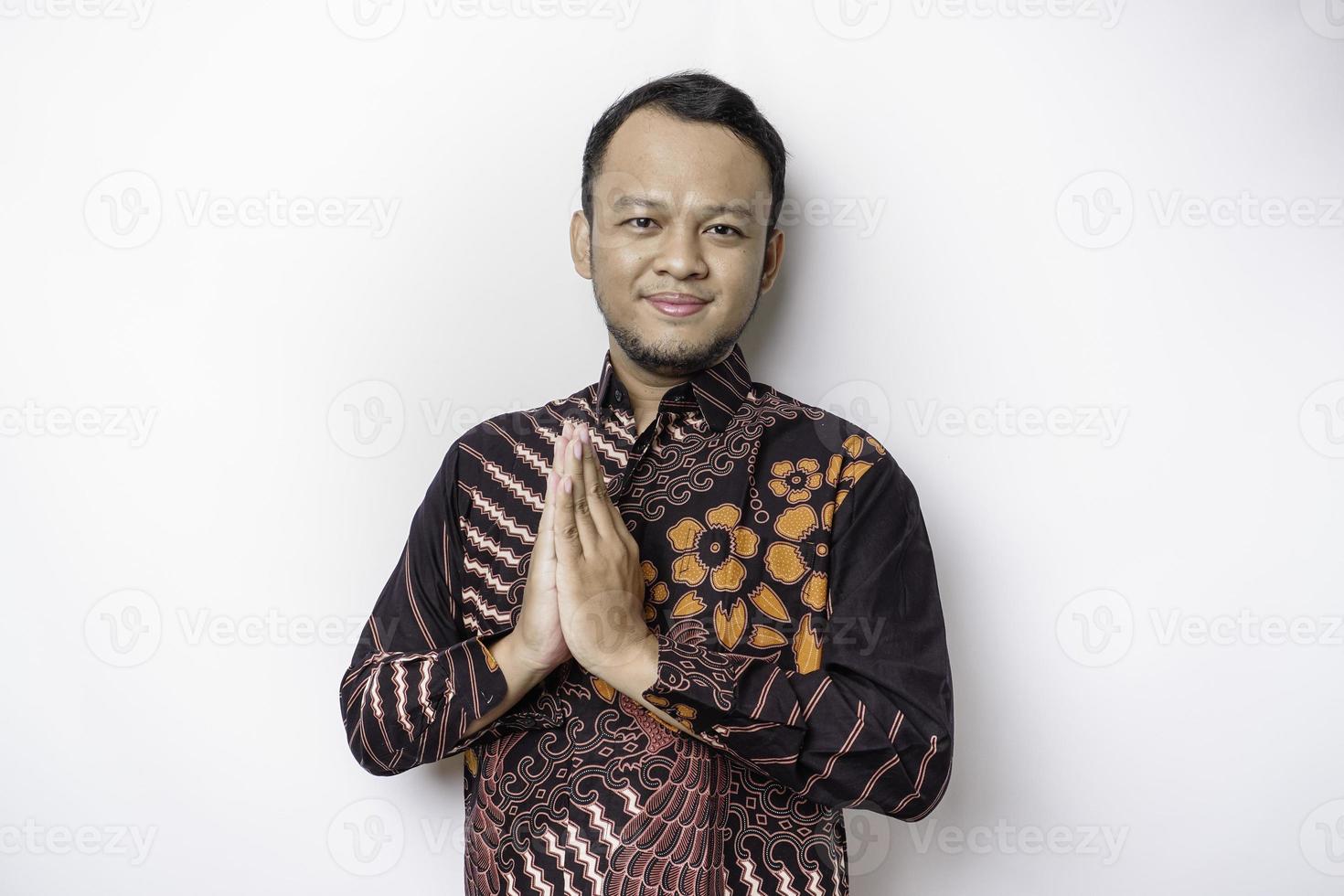 jeune homme asiatique souriant portant une chemise batik, faisant des gestes de salutation traditionnelle isolés sur fond blanc photo