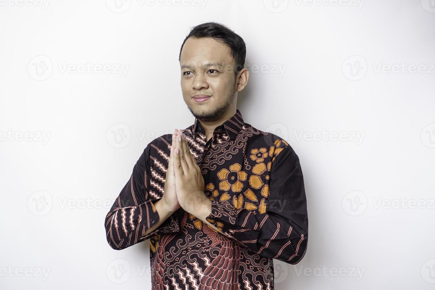 jeune homme asiatique souriant portant une chemise batik, faisant des gestes de salutation traditionnelle isolés sur fond blanc photo