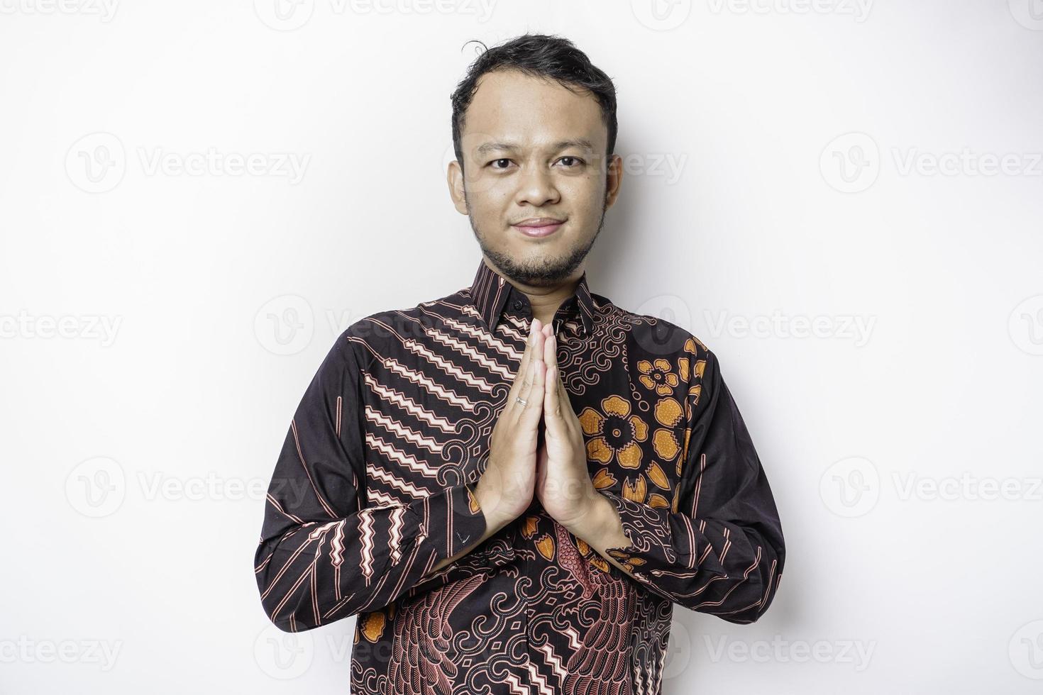 jeune homme asiatique souriant portant une chemise batik, faisant des gestes de salutation traditionnelle isolés sur fond blanc photo