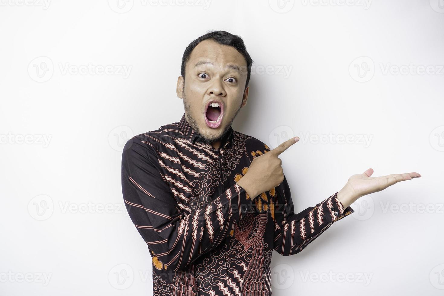 homme asiatique choqué portant une chemise batik pointant vers l'espace de copie à côté de lui, isolé sur fond blanc photo