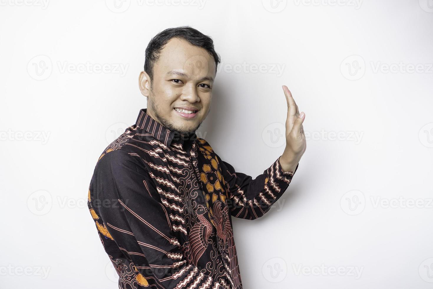 homme asiatique excité portant une chemise batik pointant vers l'espace de copie à côté de lui, isolé sur fond blanc photo