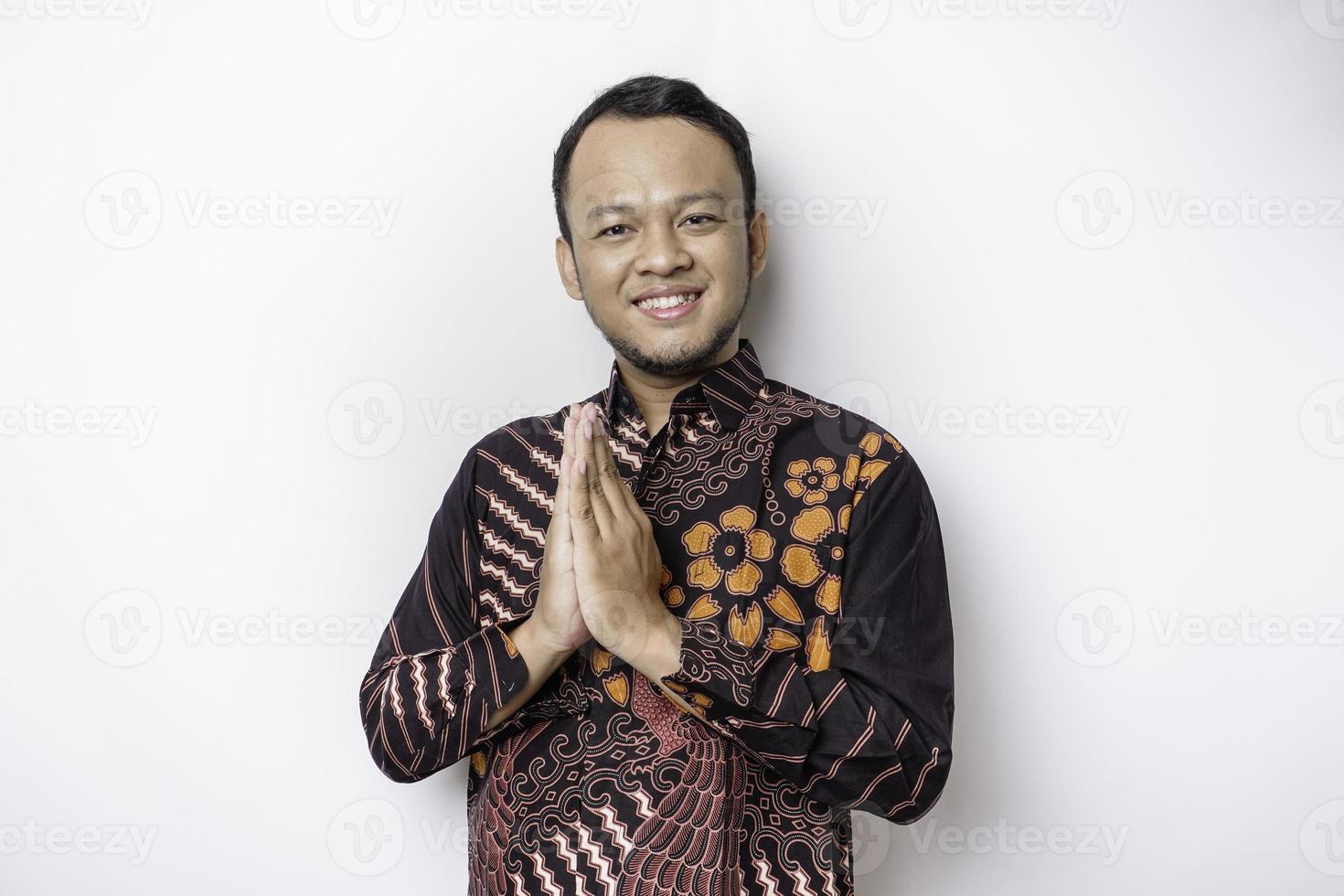 jeune homme asiatique souriant portant une chemise batik, faisant des gestes de salutation traditionnelle isolés sur fond blanc photo