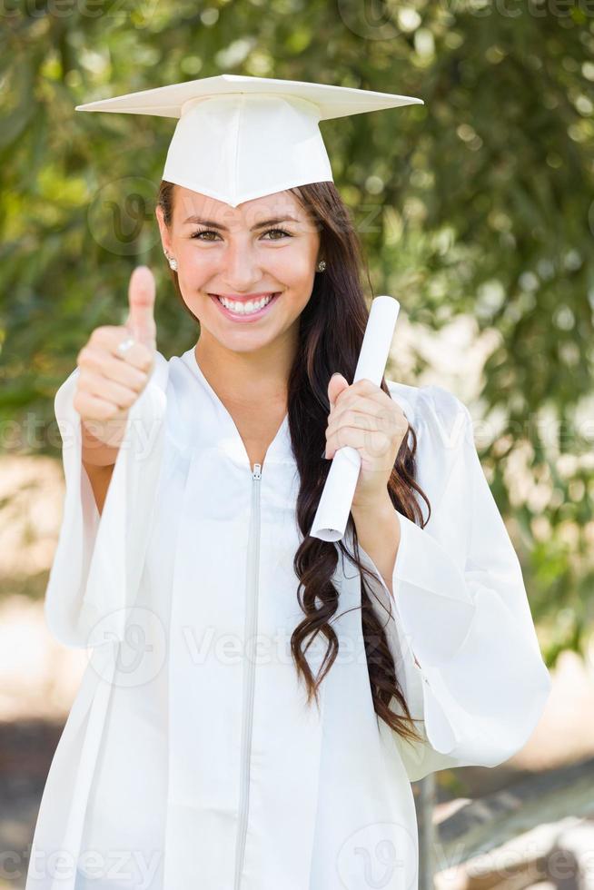 mixed race thumbs up girl célébrant l'obtention du diplôme à l'extérieur en bonnet et robe avec diplôme en main photo