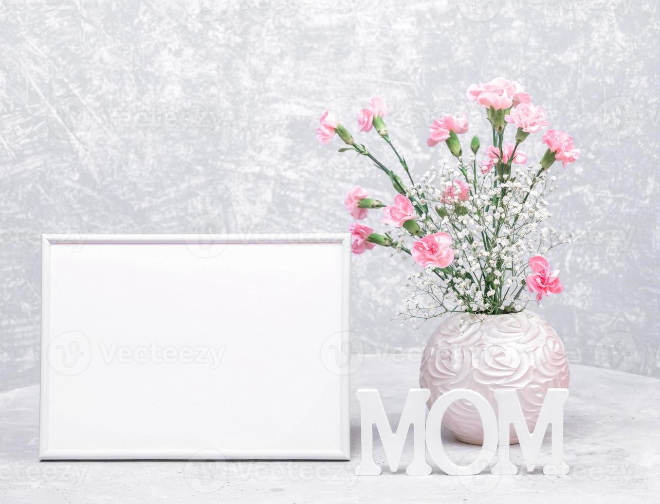 maquette cadre vide blanc, vase rond de fleurs d'oeillets et de gypsophile, lettres de maman en bois blanc. photo