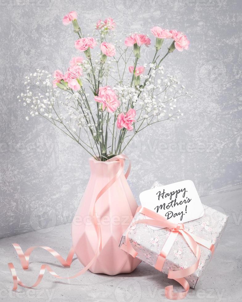 note de bonne fête des mères sur une boîte cadeau avec ruban, oeillet rose, gypsophile blanche dans un vase sur gris. photo