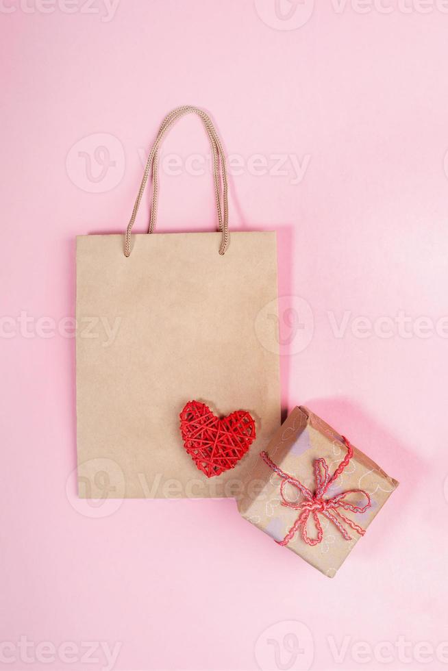 maquette de la saint-valentin avec sac en papier, boîte-cadeau emballée à la main et coeur rouge sur fond rose. concept festif zéro déchet. photo
