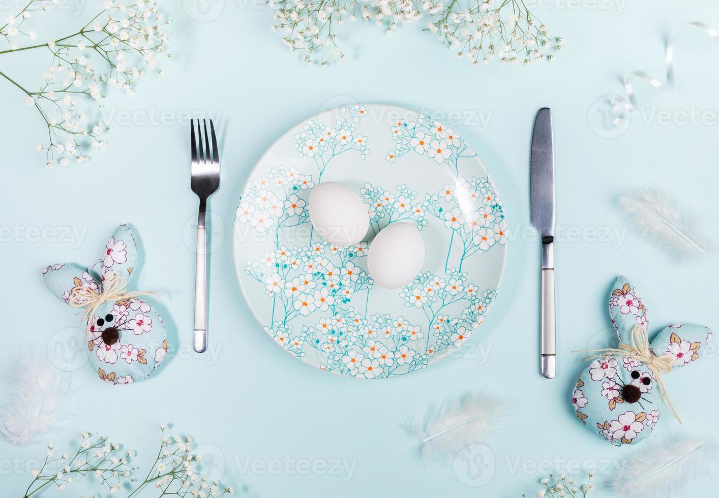 table de pâques avec des oeufs blancs sur une assiette à décor floral, des lapins de pâques et des fleurs blanches sur bleu pastel. photo