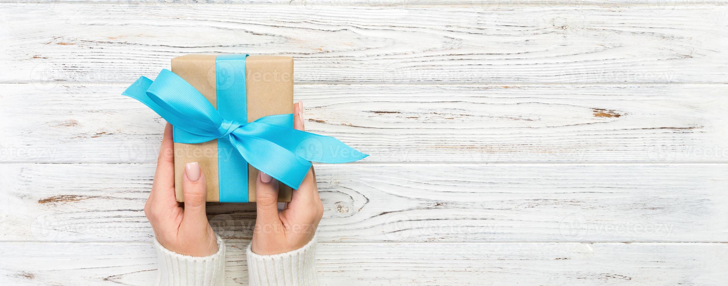 les mains de la femme donnent la saint-valentin enveloppé ou un autre cadeau de vacances fait à la main dans du papier avec un ruban bleu. boîte cadeau, décoration de cadeau sur table en bois blanc, bannière vue de dessus avec espace de copie pour votre conception photo