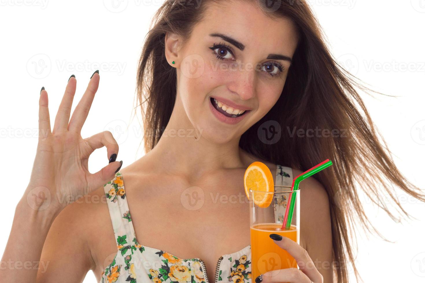 jeune fille brune heureuse en sarafan avec motif floral boit un cocktail orange souriant à la caméra et montrant ok isolé sur fond blanc photo