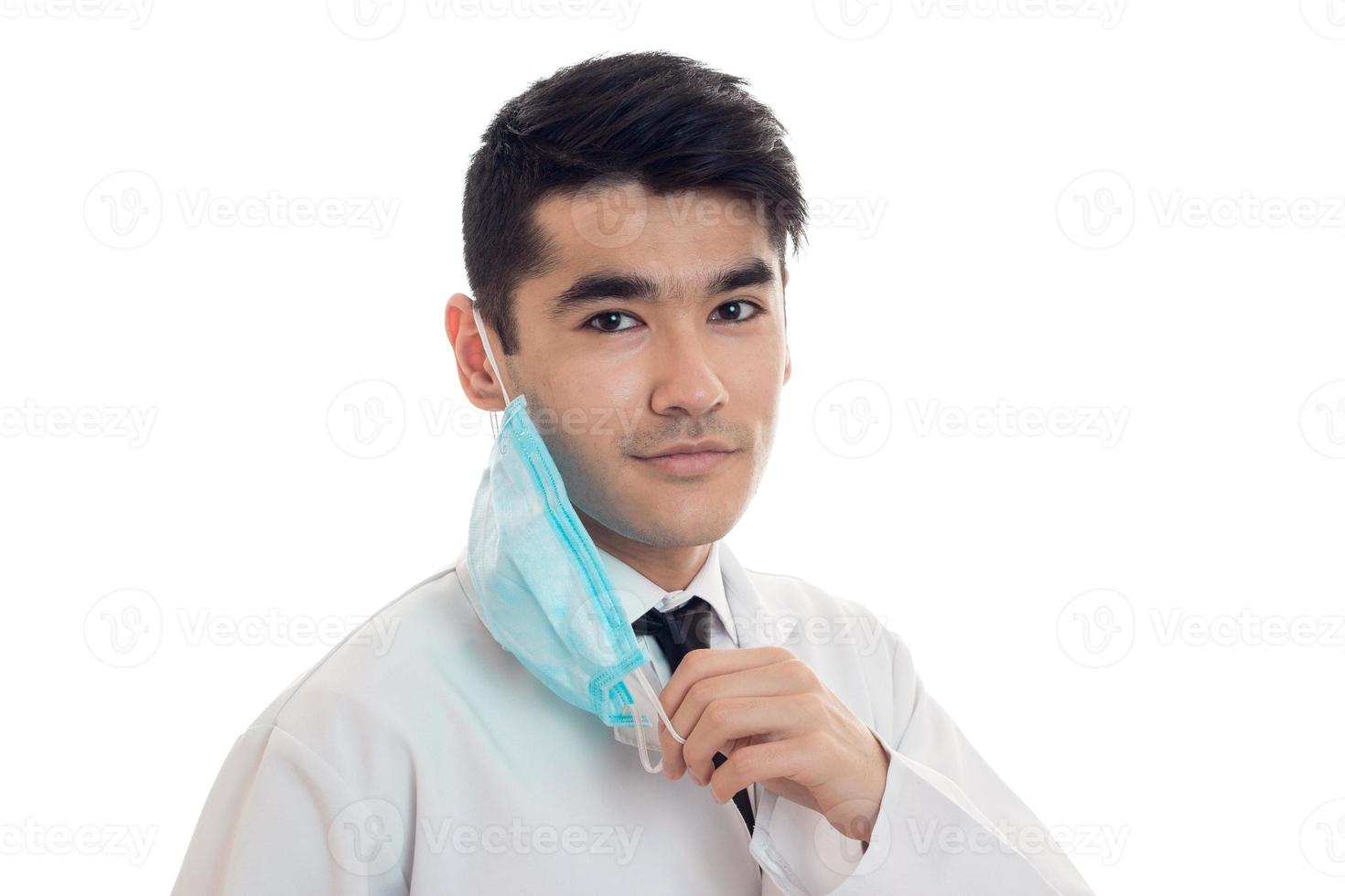 portrait d'un jeune homme brune gai médecin en uniforme blanc souriant à la caméra avec stéthoscope sur le cou et enlever un masque isolé sur fond vide photo