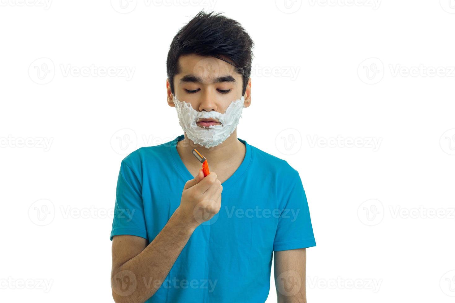 jeune homme charmant aux cheveux noirs et t-shirt bleu avec de la mousse sur le visage regardant une machine à raser photo