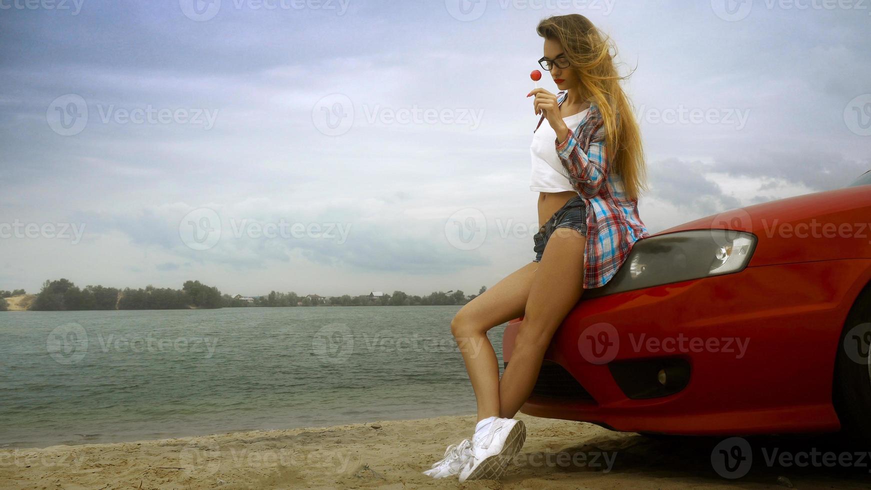 blonde sensuelle à lunettes est assise sur une voiture rouge avec une sucette dans les mains photo