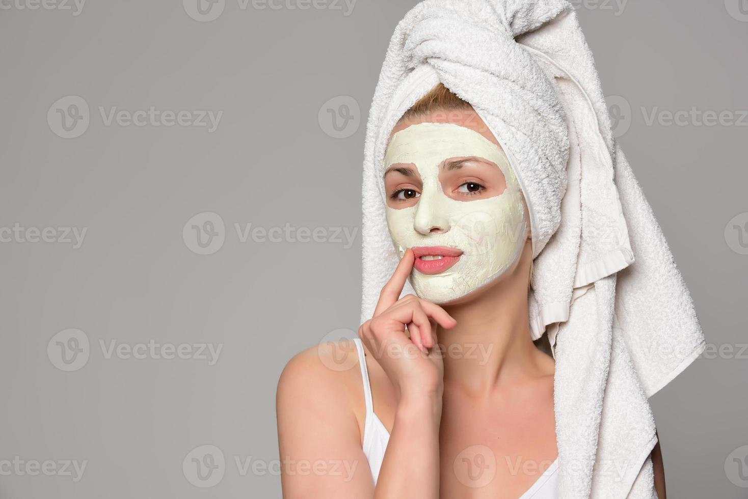 beau modèle féminin avec une serviette sur son oreille et un masque cosmétique facial blanc sur son visage. concept cosmétique de beauté. isolé sur fond gris. photo