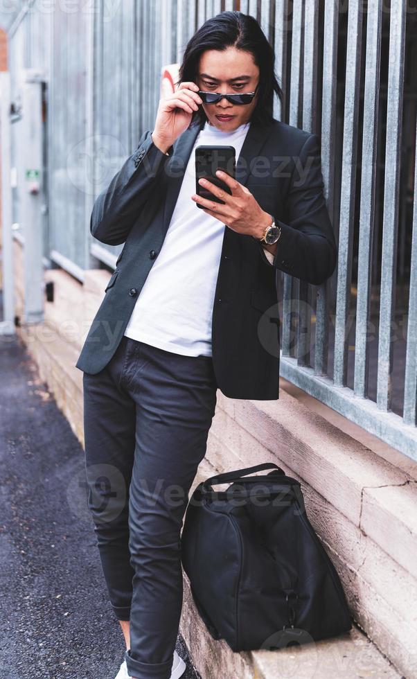 portrait d'un jeune homme asiatique beau et souriant en costume semi-formel à l'aide d'un téléphone portable photo