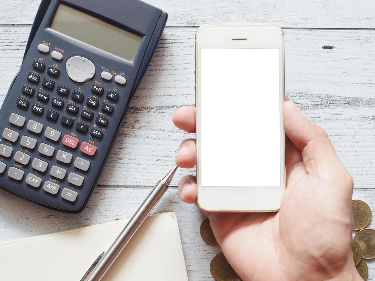 main tenant un écran blanc de smartphone avec un stylo argenté sur une calculatrice de journal de page vierge et un groupe de pièces d'or sur une table en bois blanc vue de dessus nature ombre, concept d'entreprise photo