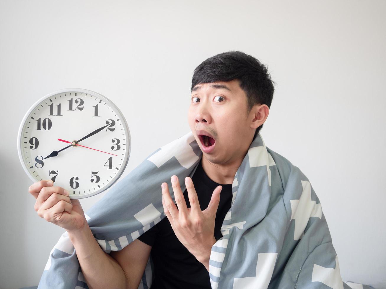 homme asiatique se réveiller avec le corps de couverture de couverture et s'ennuyer au visage regarder l'horloge à la main sur blanc isolé photo