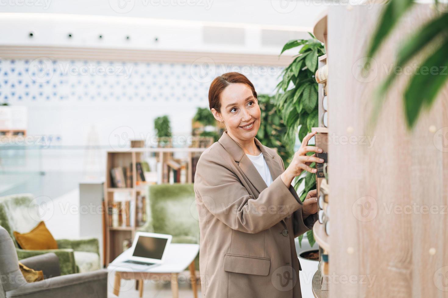 adulte femme d'affaires brune souriante de quarante ans aux cheveux longs en costume beige élégant et jeans dans un lieu public, bureau d'espace ouvert vert, coworking. enseignant ou mentor sympathique avec un livre à la bibliothèque photo