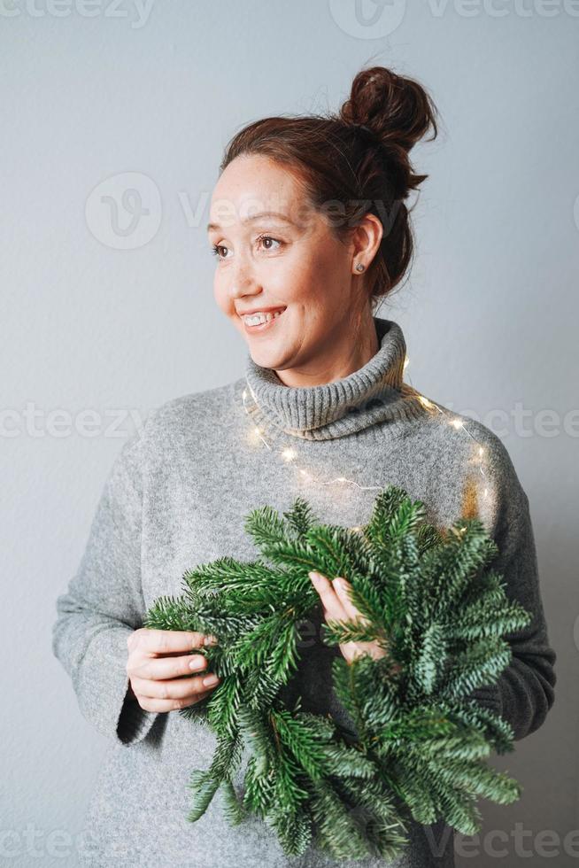 adulte belle femme souriante de quarante ans avec des cheveux bouclés brune en robe tricotée gris chaud avec une guirlande de noël sapin bricolage à la main sur le fond gris photo