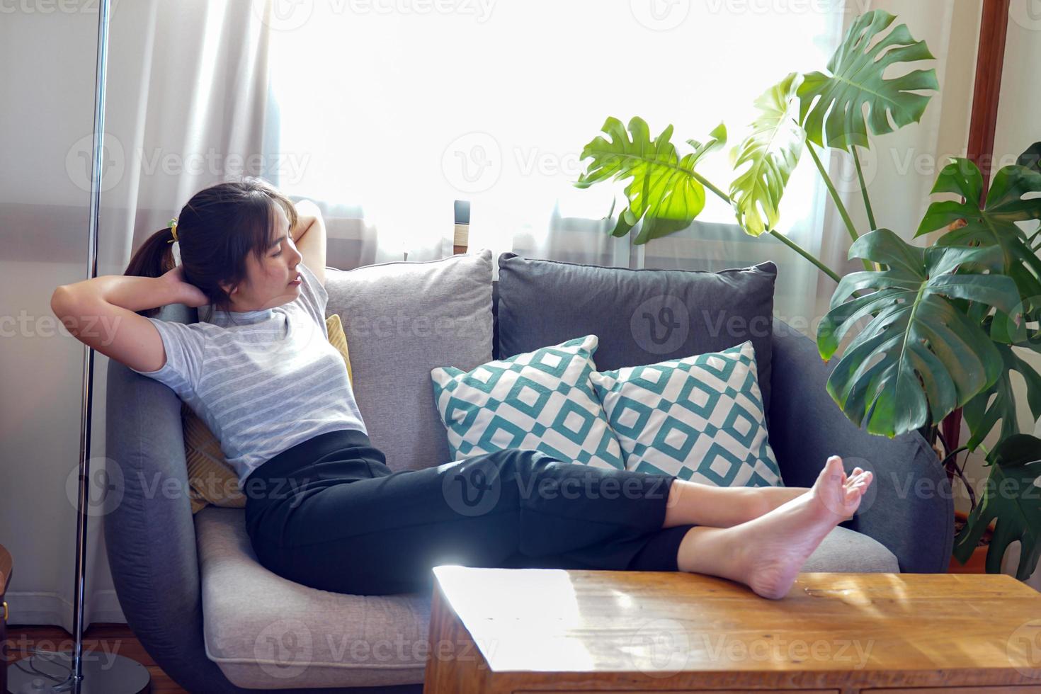 une femme asiatique s'allonge et se détend sur le canapé, elle pose ses jambes contre la table. mets ta tête sous les mains entrelacées. concept se détendre après le travail, se détendre, coin confortable, coin calme, faire une sieste photo