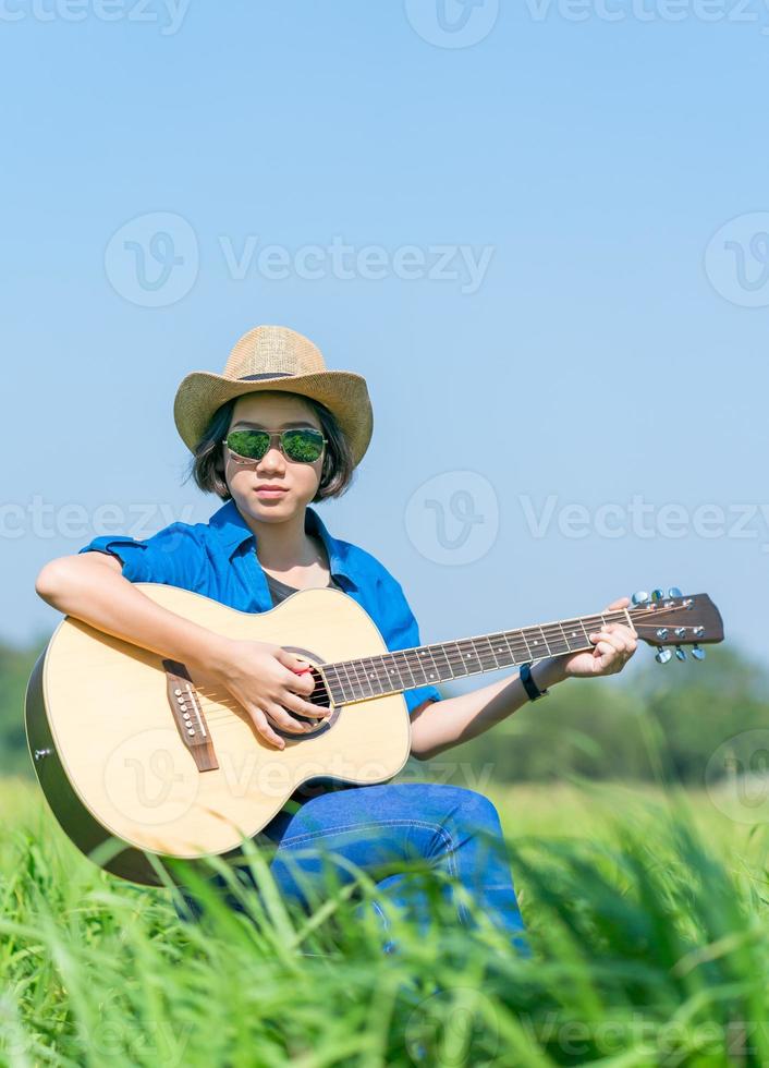 les femmes cheveux courts portent un chapeau et des lunettes de soleil s'assoient en jouant de la guitare dans le champ d'herbe photo