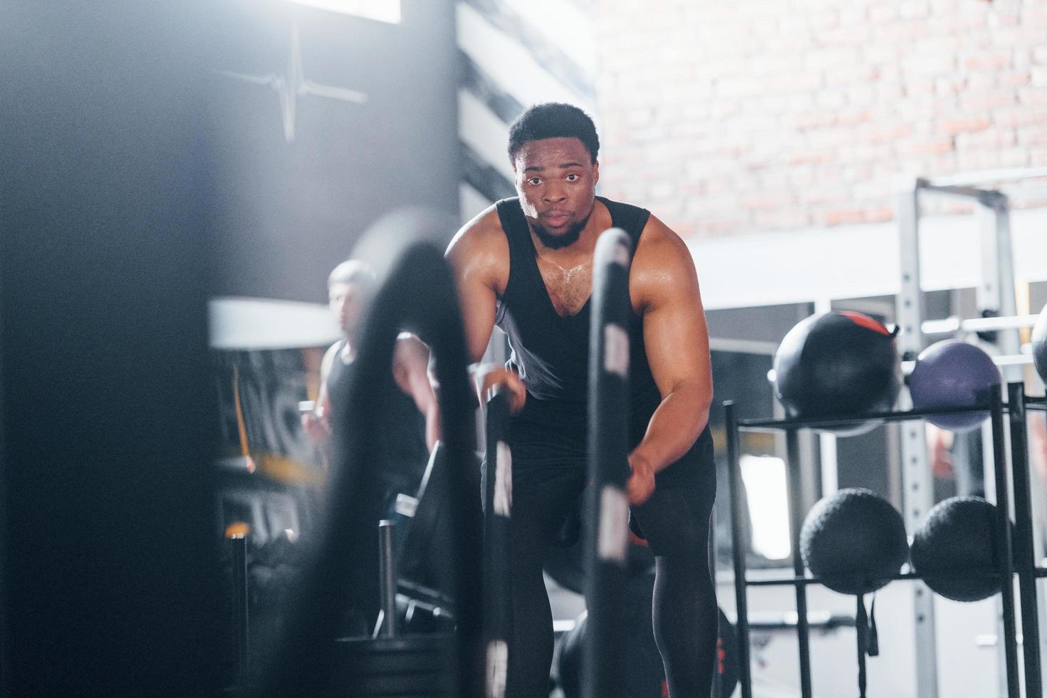 un homme afro-américain fort en vêtements sportifs a une journée d'entraînement dans la salle de sport. faire du crossfit en utilisant des cordes photo