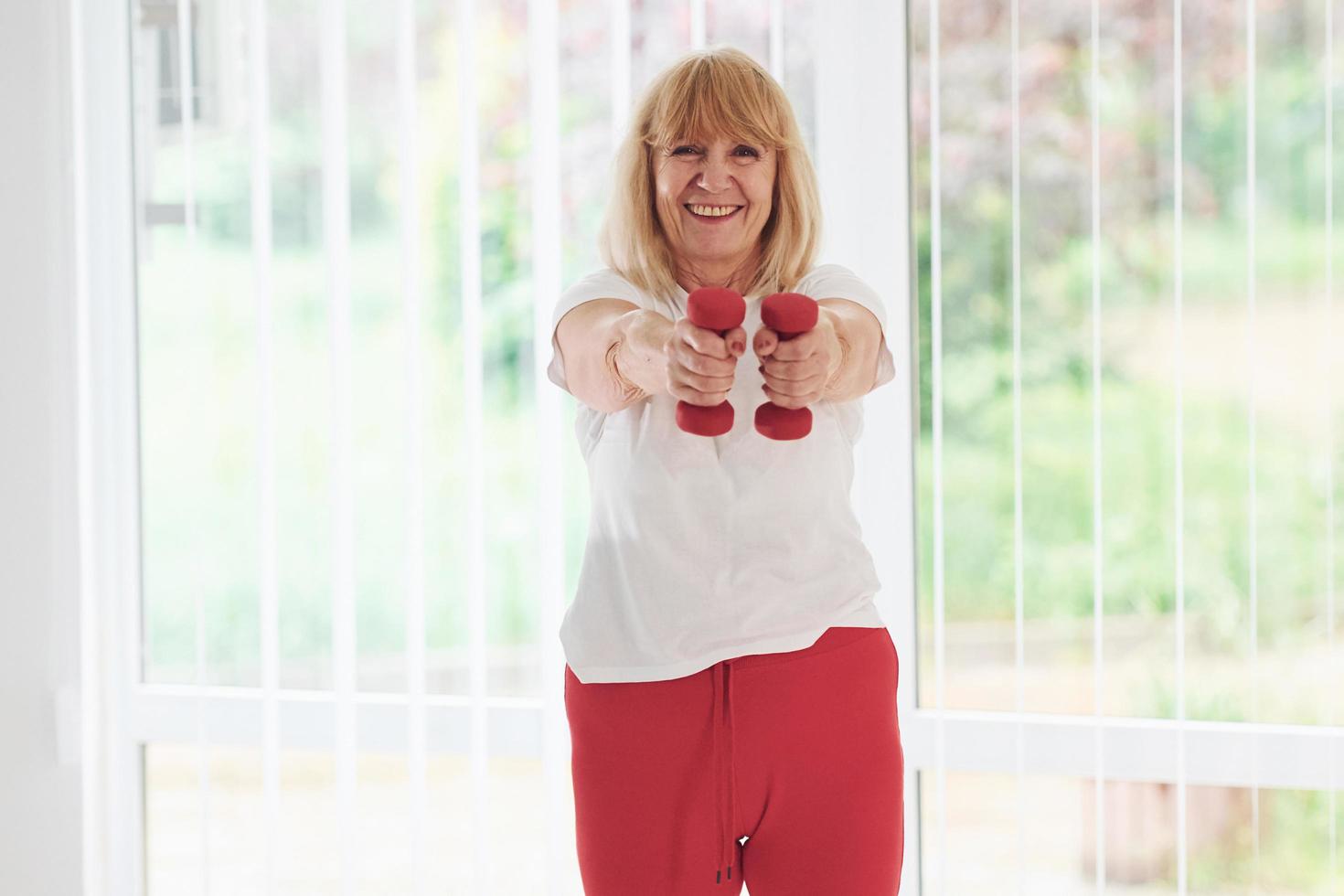 femme senior positive en vêtements sportifs à l'intérieur pendant la journée, faire des exercices avec des haltères photo