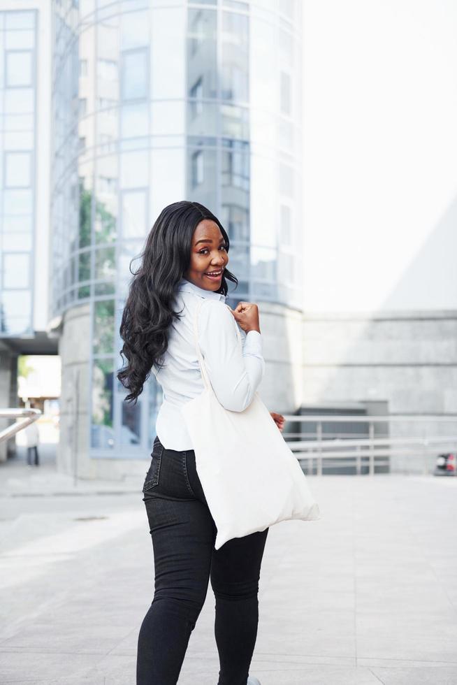 avec sac à provisions. jeune femme afro-américaine en chemise blanche à l'extérieur de la ville contre le bâtiment de l'entreprise photo