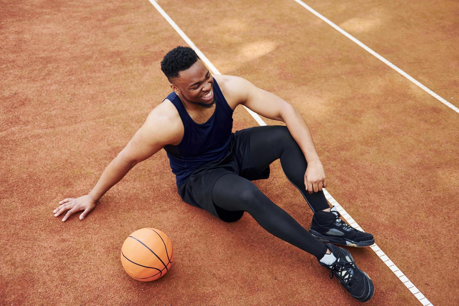 un homme afro-américain fatigué est assis sur le sol avec un ballon sur le terrain à l'extérieur. fait une pause photo