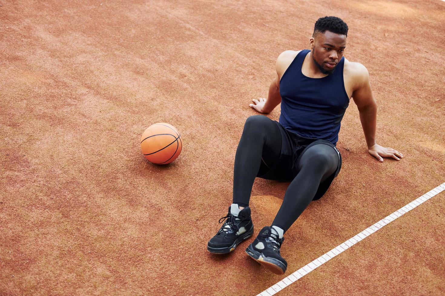 un homme afro-américain fatigué est assis sur le sol avec un ballon sur le terrain à l'extérieur. fait une pause photo