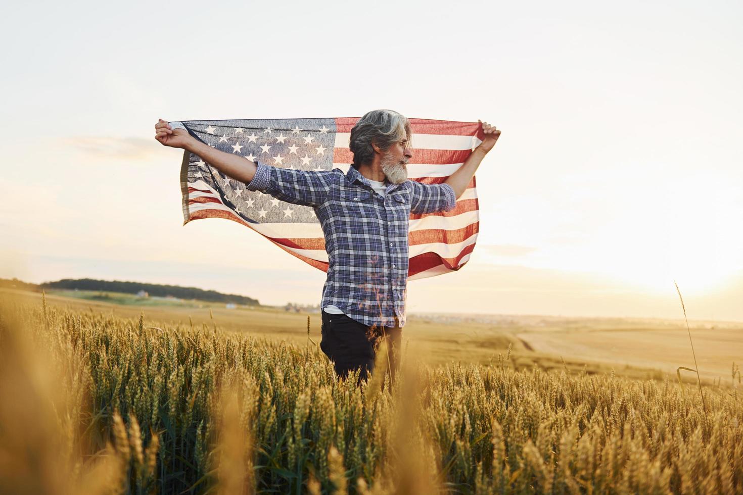 tenant le drapeau américain dans les mains. homme élégant senior patriotique aux cheveux gris et à la barbe sur le terrain agricole photo