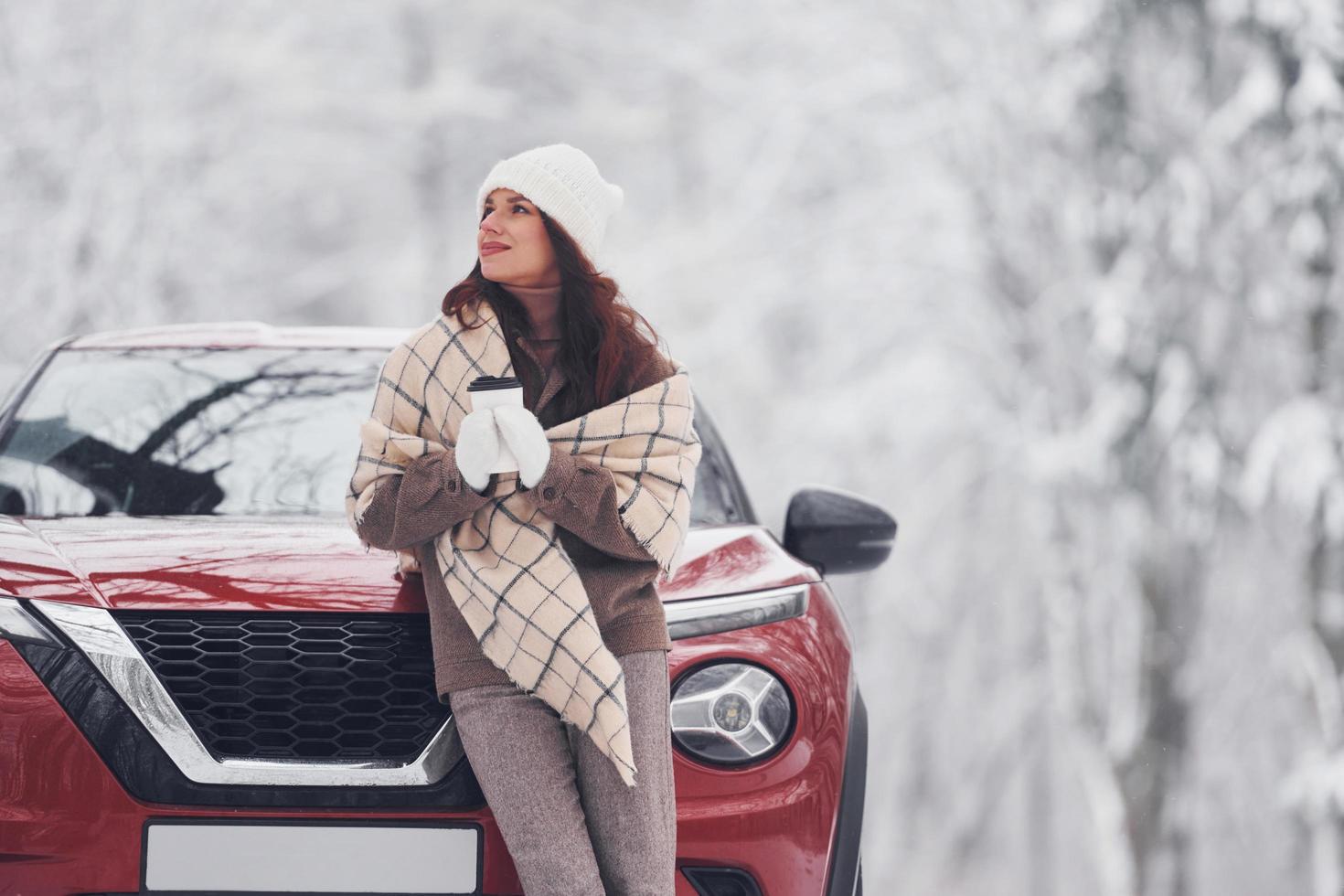 devant sa voiture. belle jeune femme est à l'extérieur près de son automobile rouge à l'heure d'hiver photo